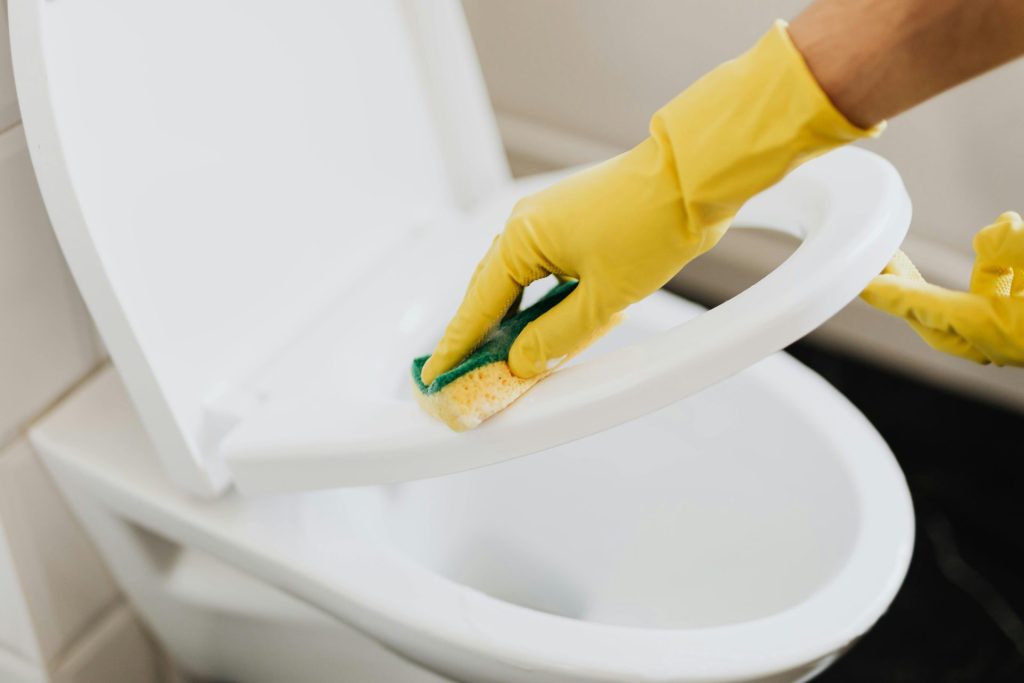 A person using yellow gloves and a sponge to clean a toilet seat indoors.